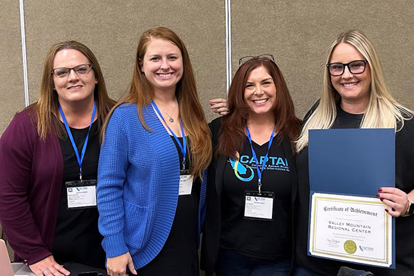 Four women stand indoors, smiling at the camera; one holds a Certificate of Achievement from Valley Mountain Regional Center.