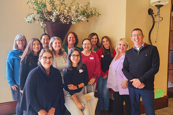 A group of twelve adults stand indoors, smiling for a photo in front of a beige wall with a large floral arrangement.