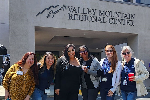Six women stand smiling in front of the Valley Mountain Regional Center building. Four wear badges and lanyards, one holds a red cup, and the group poses outdoors in daylight.
