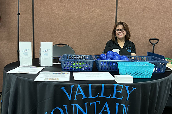 A woman sits at a booth table with brochures, papers, and baskets of blue stress balls. The tablecloth reads 