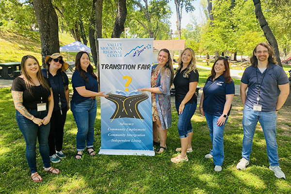 Seven people stand outdoors around a “Transition Fair” banner for the Valley Mountain Regional Center, smiling at the camera in a park setting with trees and grass.