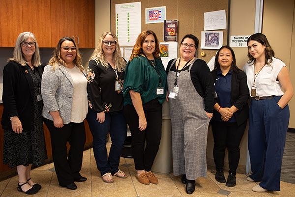 Seven women stand indoors in an office space, posing together and smiling for a group photo.