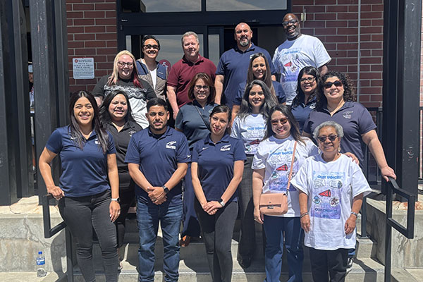 A group of people pose for a photo on outdoor steps in front of a brick building, wearing matching shirts and smiling at the camera.