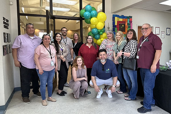 A group of twelve adults pose together indoors near a display of green and yellow balloons, smiling at the camera.