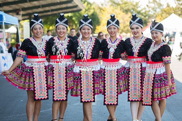 Six women stand in a row wearing traditional Hmong dresses with colorful patterns, silver accessories, and headdresses at an outdoor event.