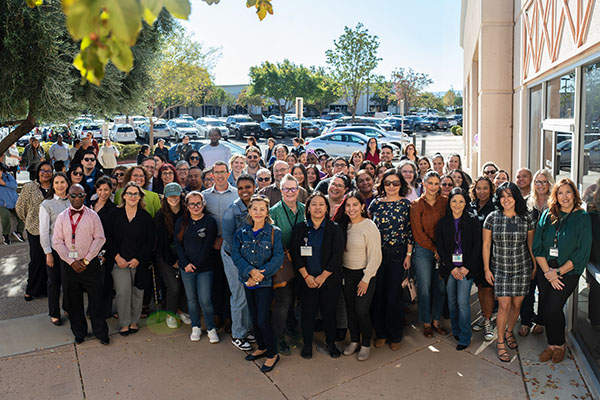 A large group of adults poses for a group photo outdoors near a building on a sunny day, with cars and trees visible in the background.