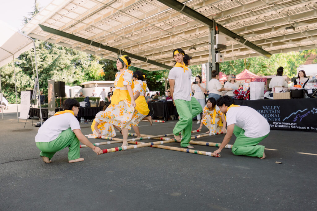 Teenagers performing the traditional Filipino Tinikling dance at VMRC Cultural & Disability Fair, stepping rhythmically between bamboo poles while wearing colorful attire and smiling.