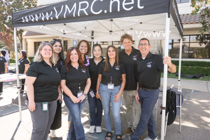 A group of seven people in black polo shirts standing together under a branded tent for VMRC.net.
